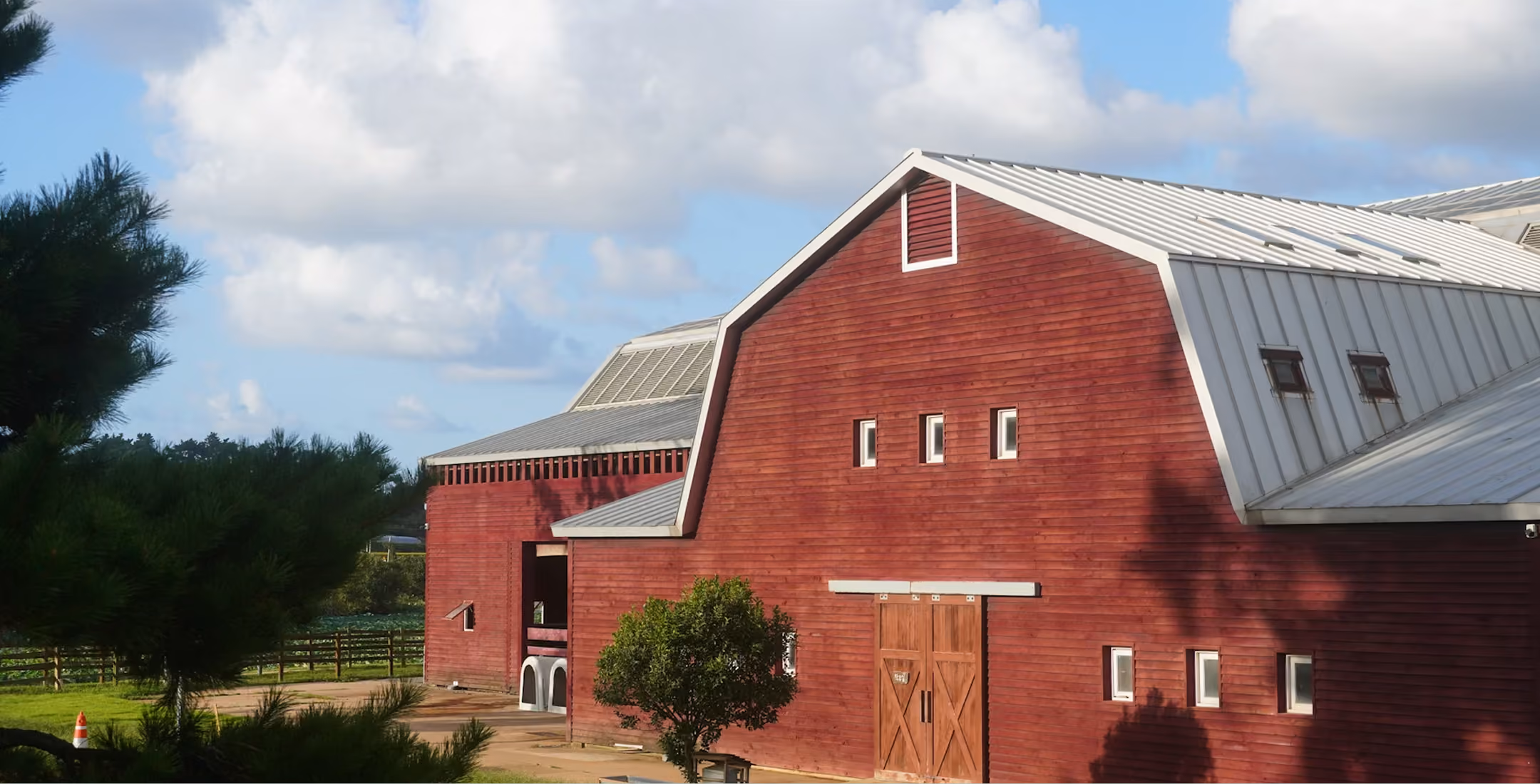 Large red barn with white metal roof and small windows amid green trees and blue sky.
