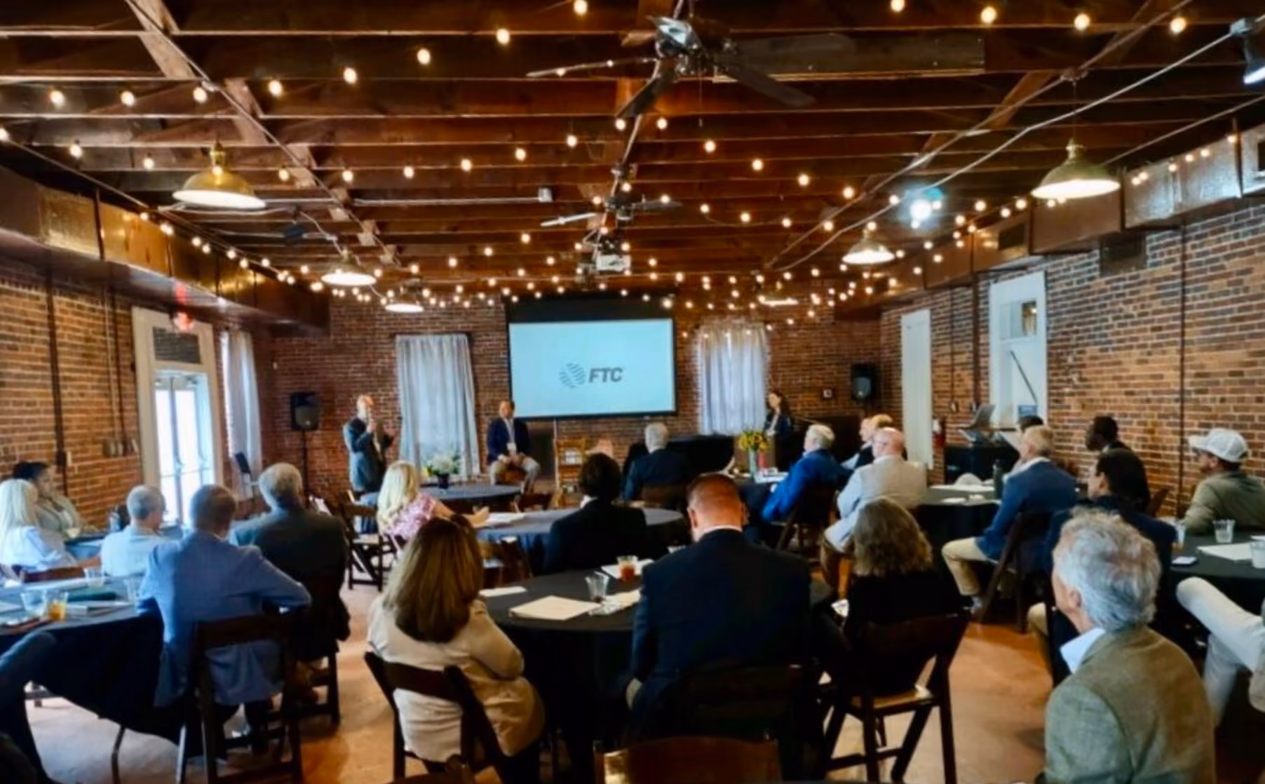People seated at round tables listening to a speaker in a string-light decorated brick room with FTC logo on screen.