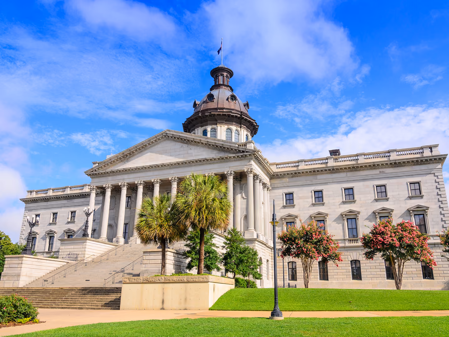 Classical government building with steps, columns, dome, palm trees, and flowering trees under blue sky.