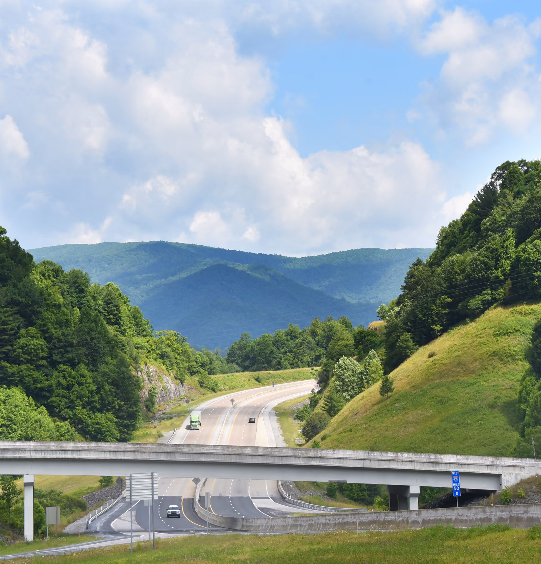 Highway with a concrete overpass surrounded by green hills and mountains under a partly cloudy sky.