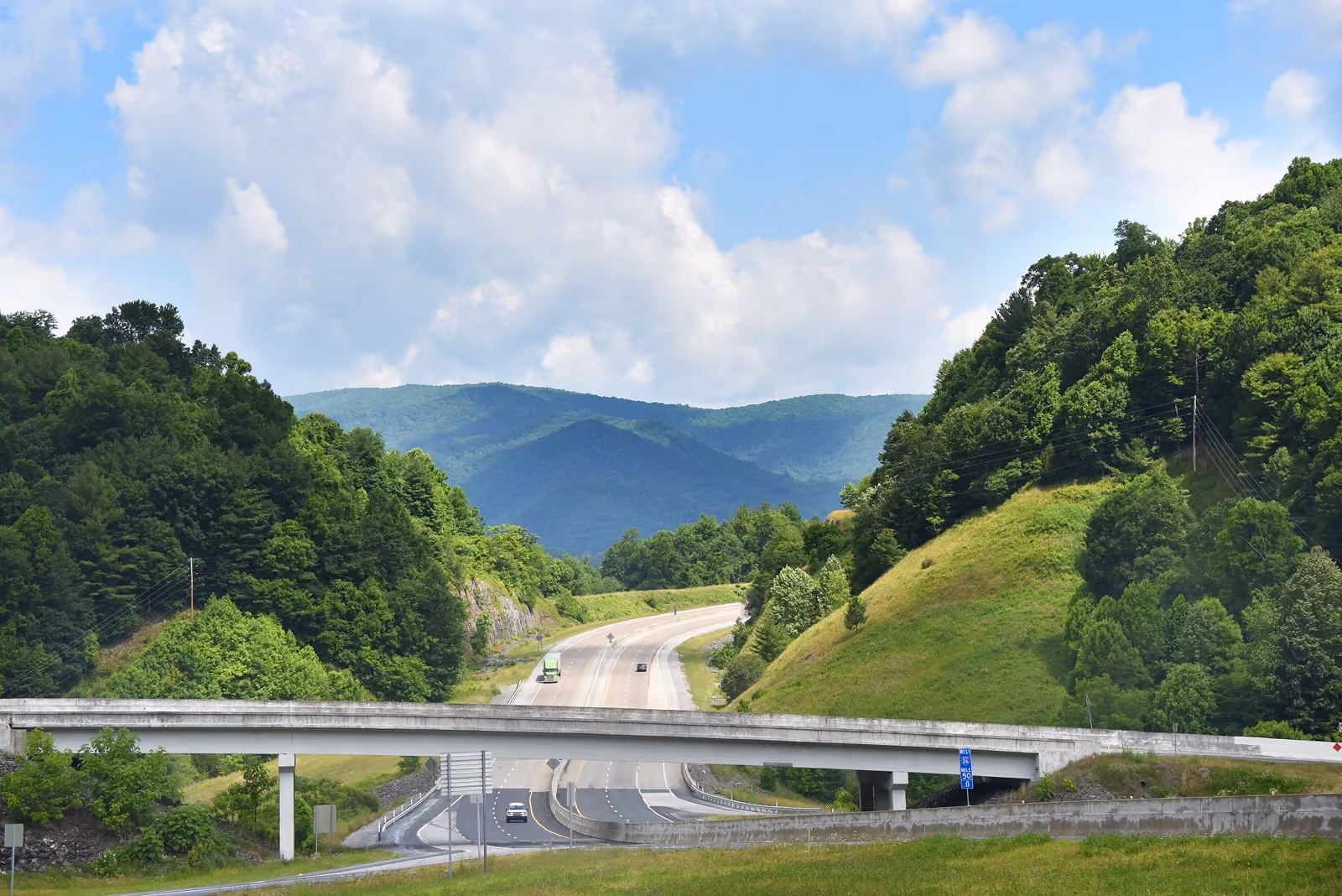 Highway passing through green forested hills with a concrete overpass and a blue sky with clouds.