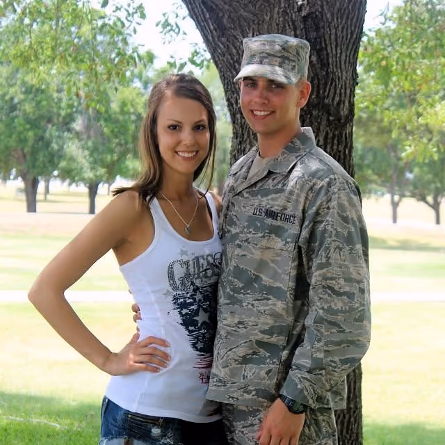 Young woman in casual clothes stands next to a smiling man in U.S. Air Force uniform under a tree.