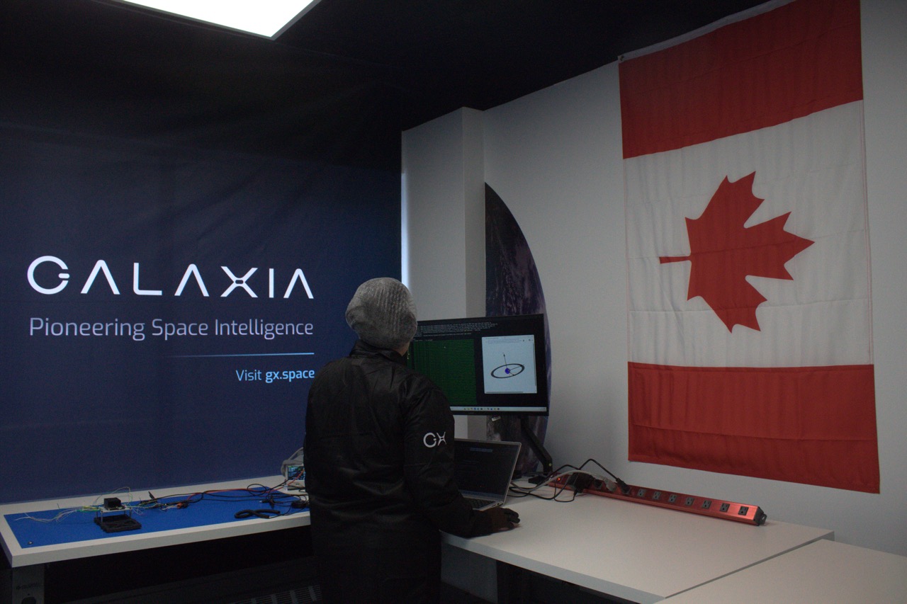 Person in a beanie and jacket works on a computer in a room with a Canadian flag and Galaxia space banner.