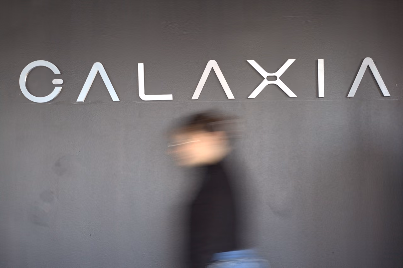 Blurred person walking past a gray wall with large silver letters spelling GALAXIA.
