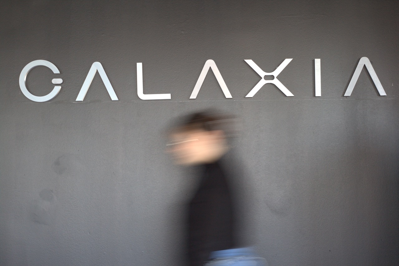 Blurred person walking past a gray wall with large silver letters spelling GALAXIA.