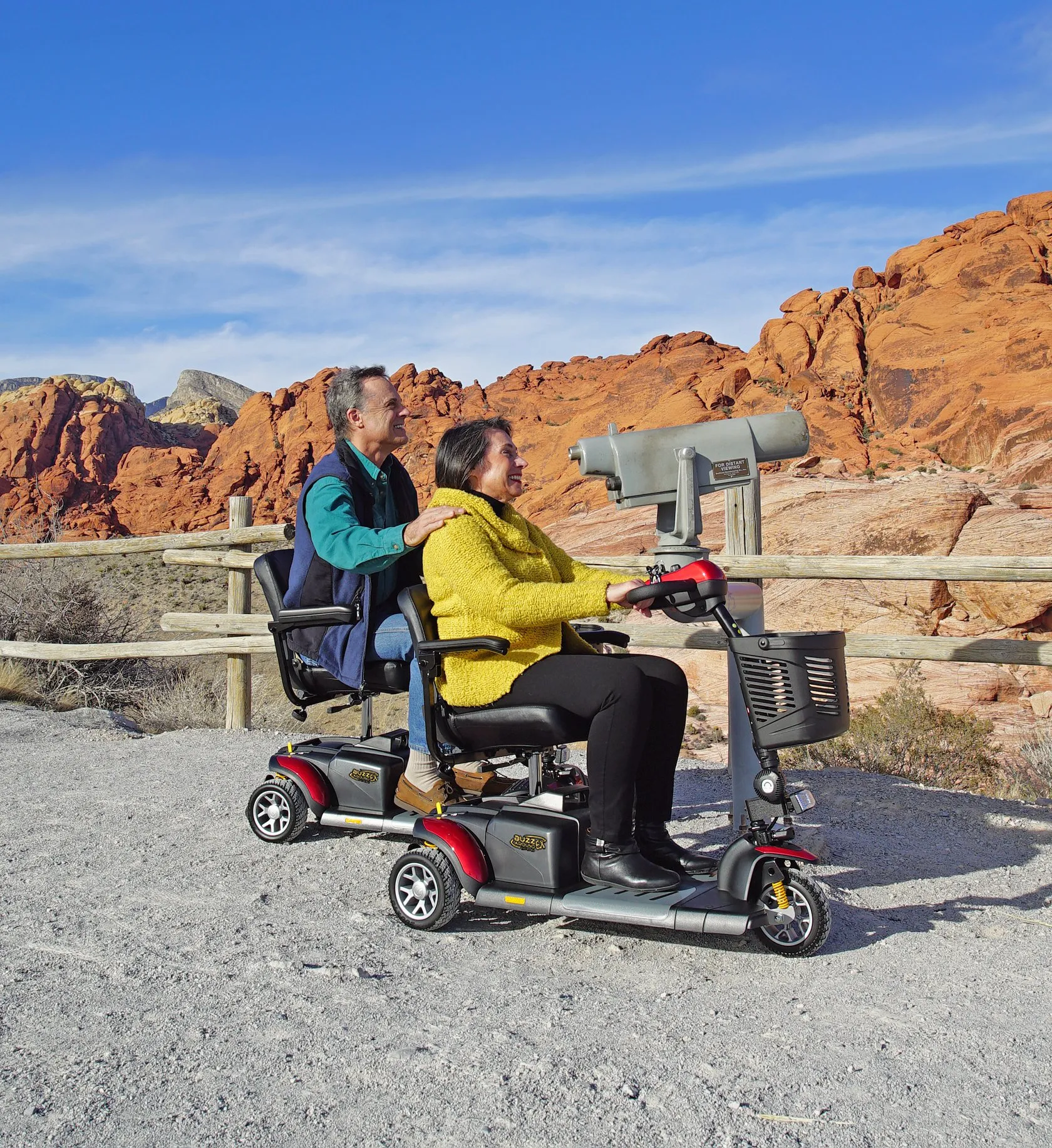 Smiling man and woman riding two red mobility scooters by a wooden fence with red rock formations behind.