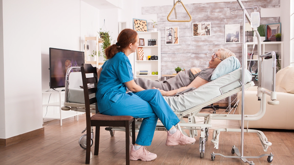 Nurse in blue scrubs sitting beside elderly woman lying in a hospital bed at home.
