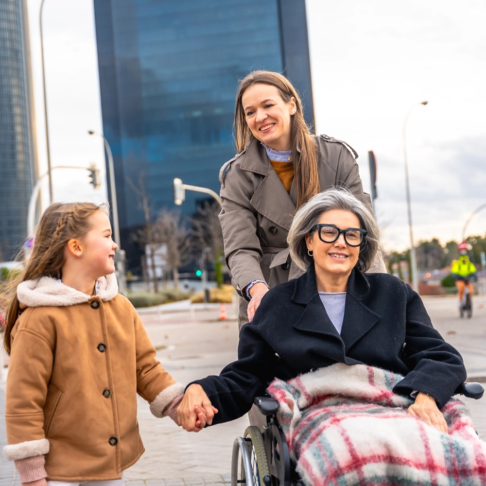 Smiling woman pushing elderly woman in wheelchair holding hands with a young girl outdoors in city.