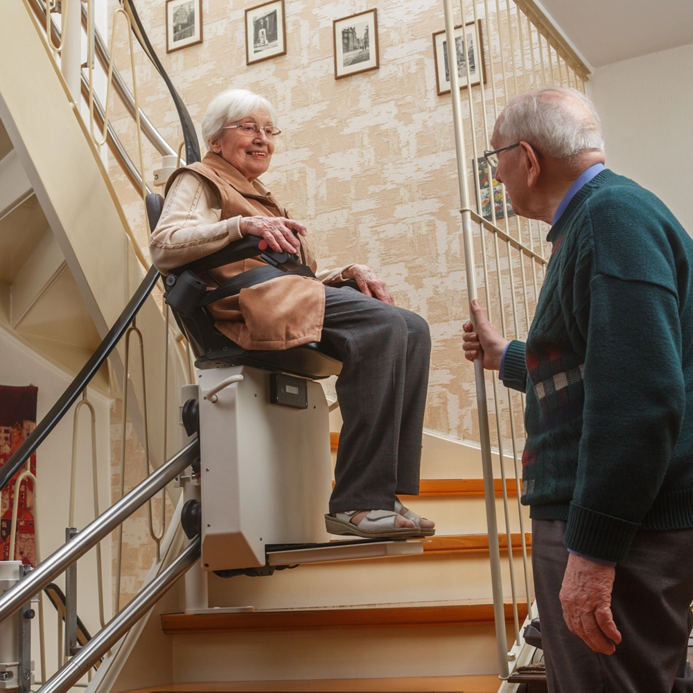 An elderly woman rides a stair lift while a man looks on at the bottom of the stairs inside a home.