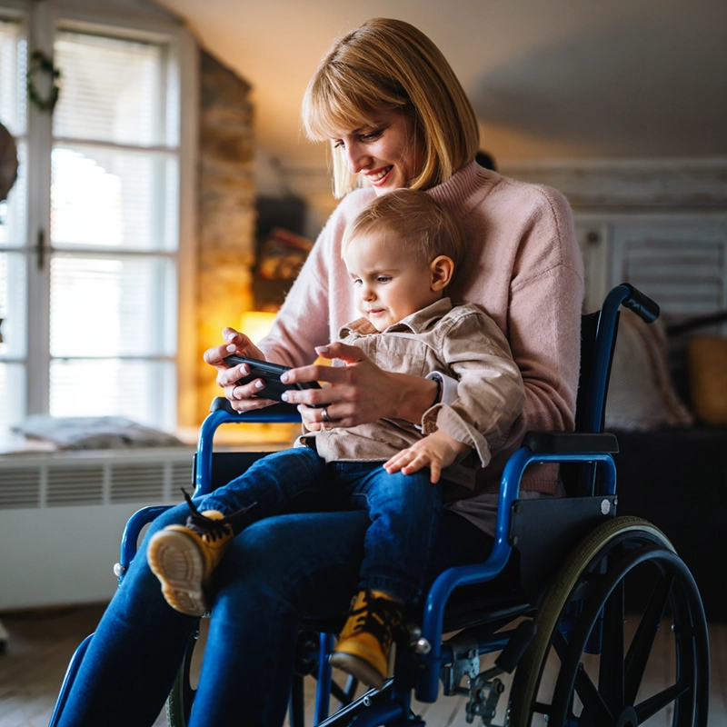 Smiling woman in wheelchair holding a toddler both looking at a smartphone together indoors.