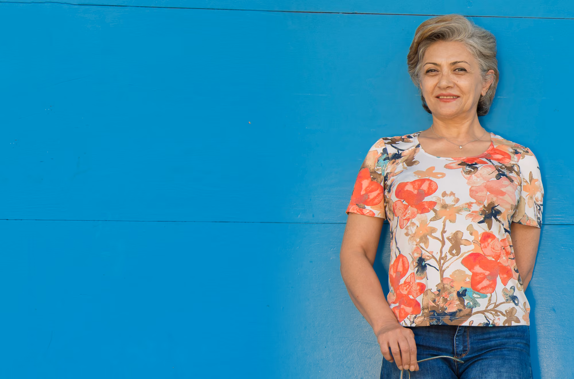 Woman in a floral top standing against a blue wall, smiling.