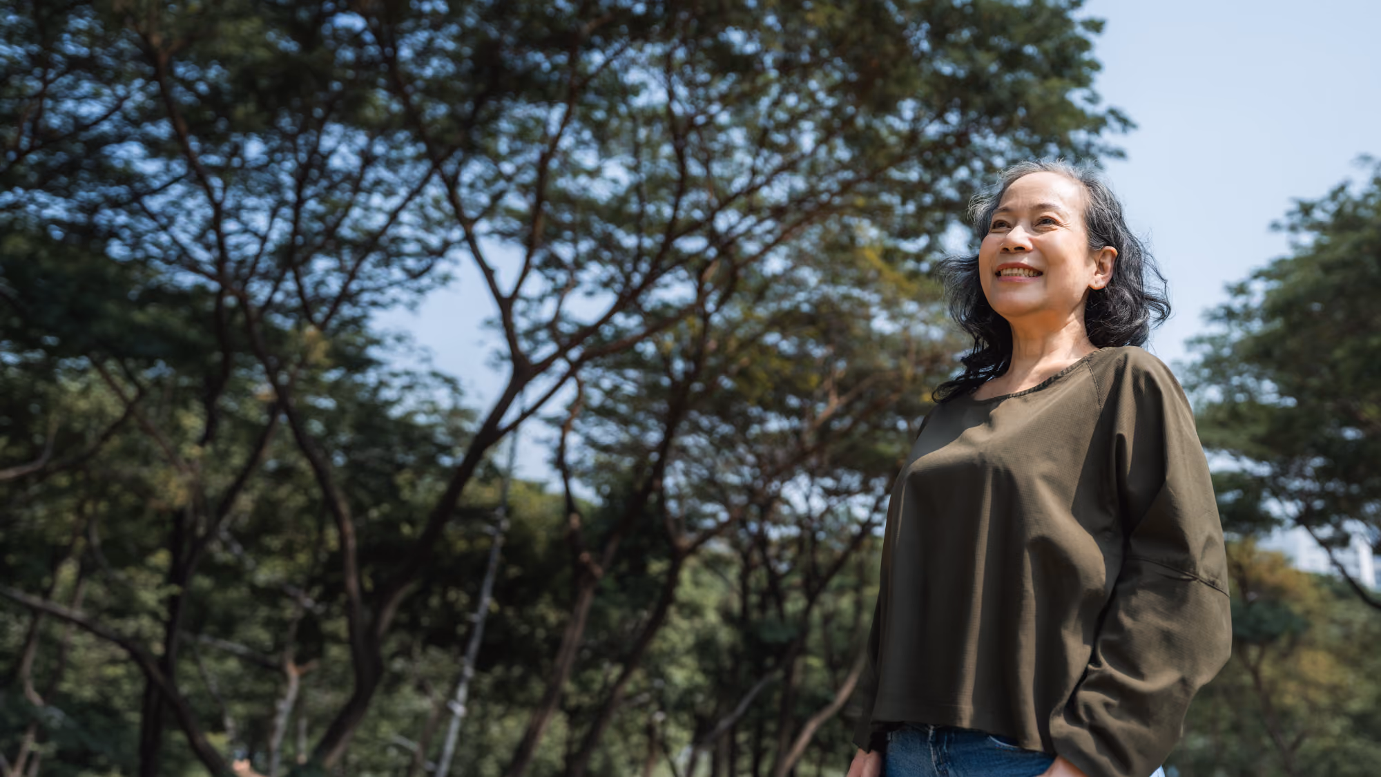 Older woman standing outdoors among trees, smiling and looking into the distance.