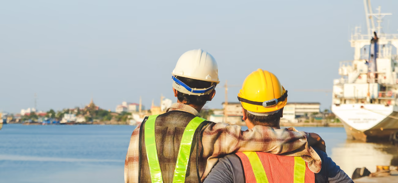 Two construction workers in hard hats and safety vests looking out over the water.