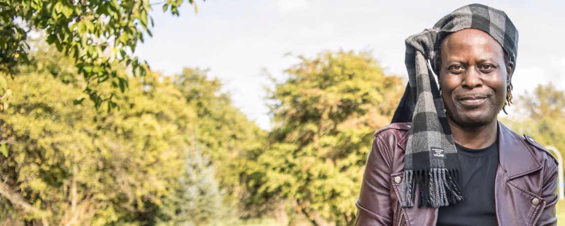 Man wearing a plaid scarf on his head standing outdoors among trees, smiling.