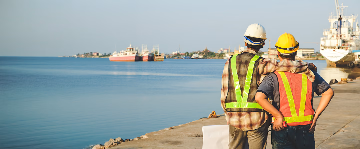 Two construction workers in hard hats and safety vests looking out over the water.