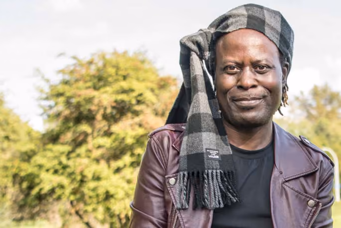 Man wearing a plaid scarf on his head standing outdoors among trees, smiling