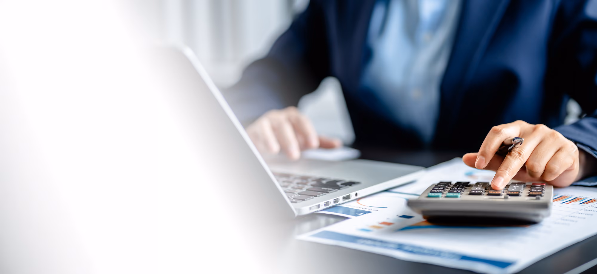 Hands using a calculator beside a laptop and printed financial charts.