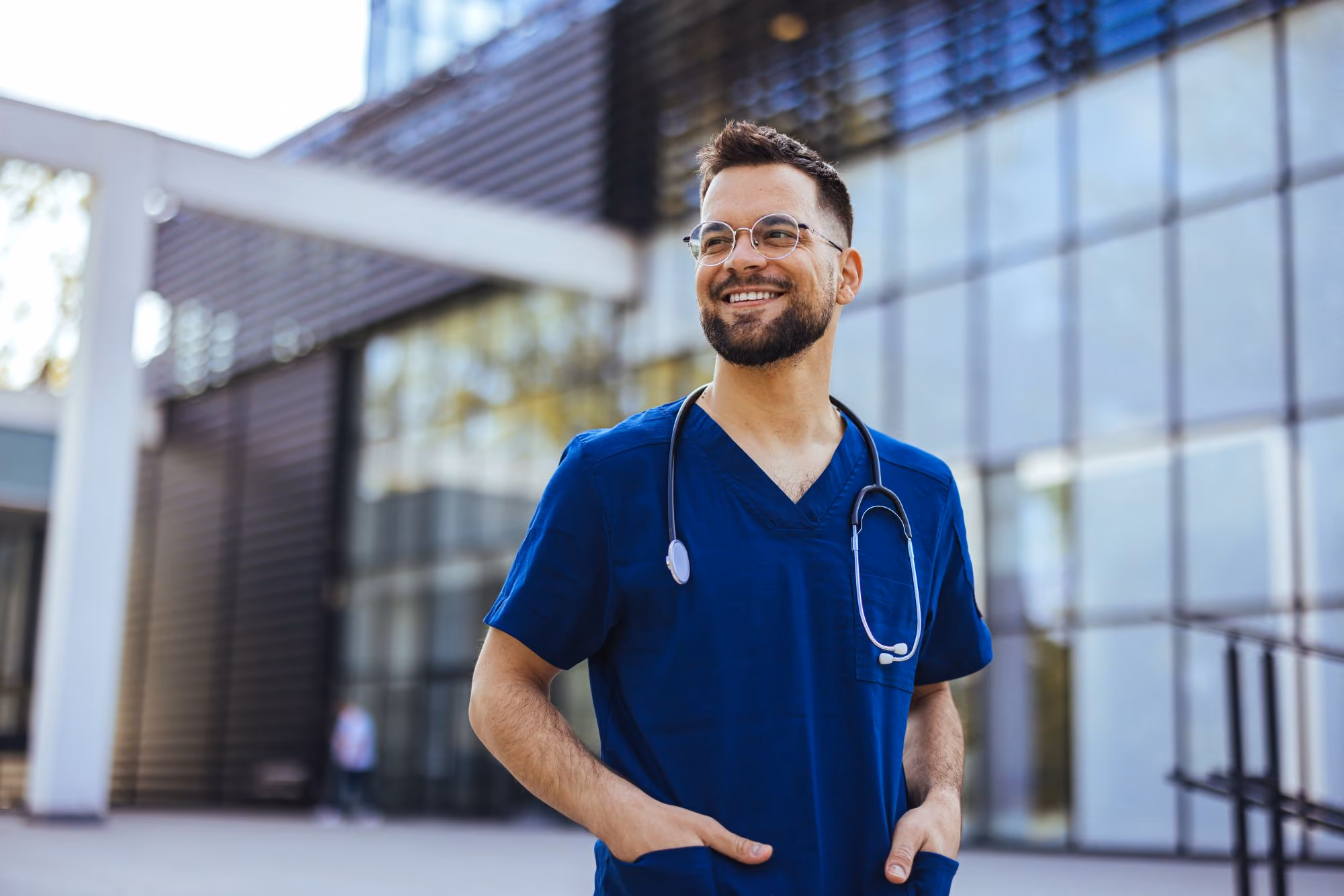 Health care provider in scrubs with a stethoscope standing outside a building, smiling.