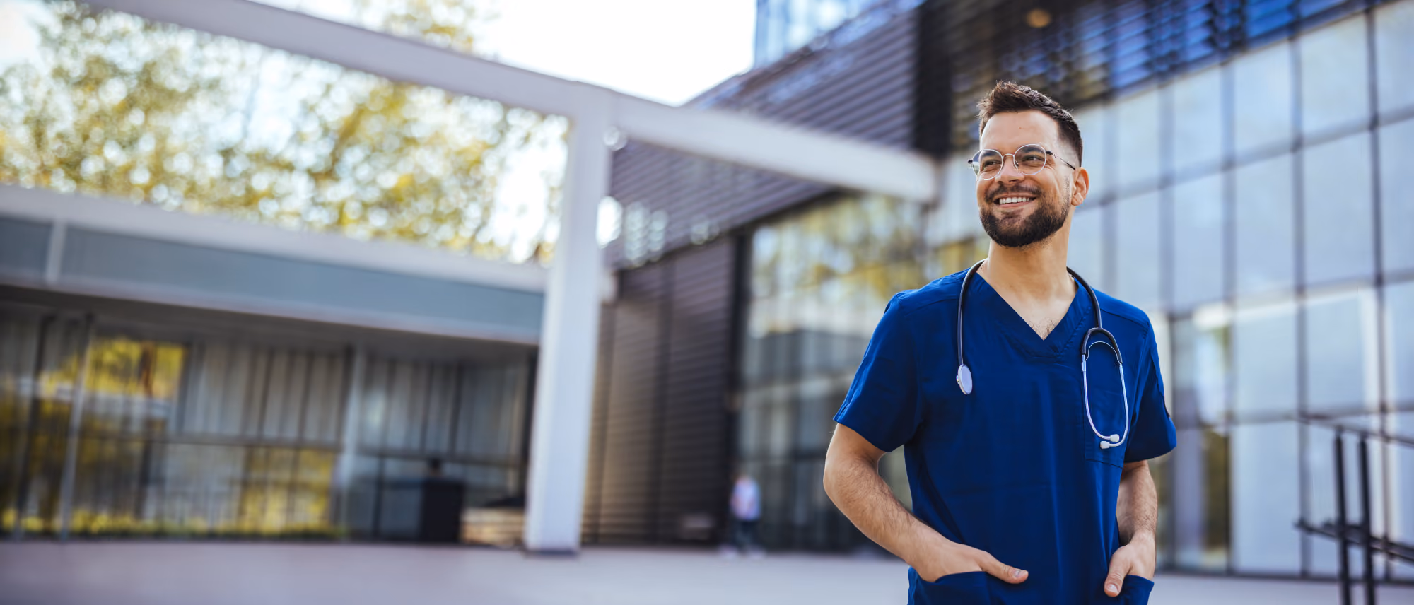 Health care provider in scrubs with a stethoscope standing outside a building, smiling.