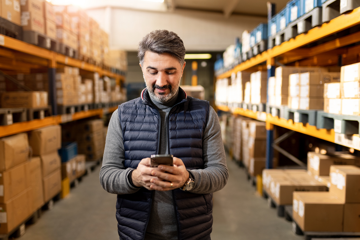 A business owner stands in his warehouse full of boxes looking at his phone in his hands smiling.