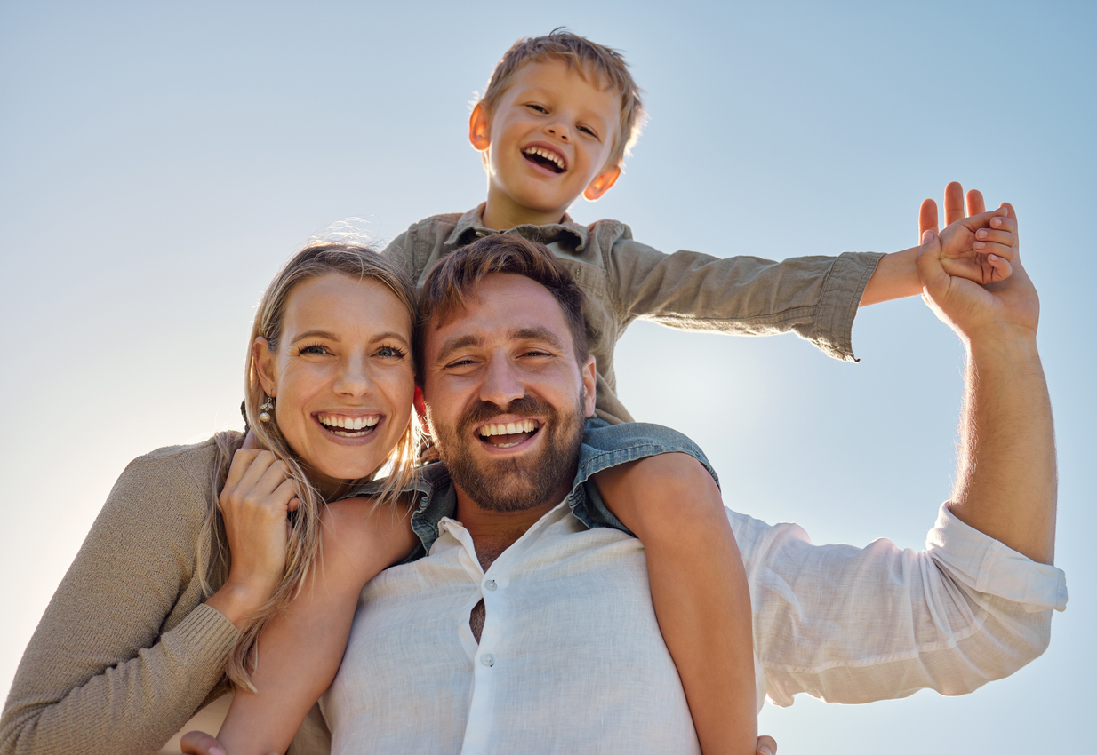 A mother and father with his son on his shoulders smile down at the camera with a bright sky behind them.