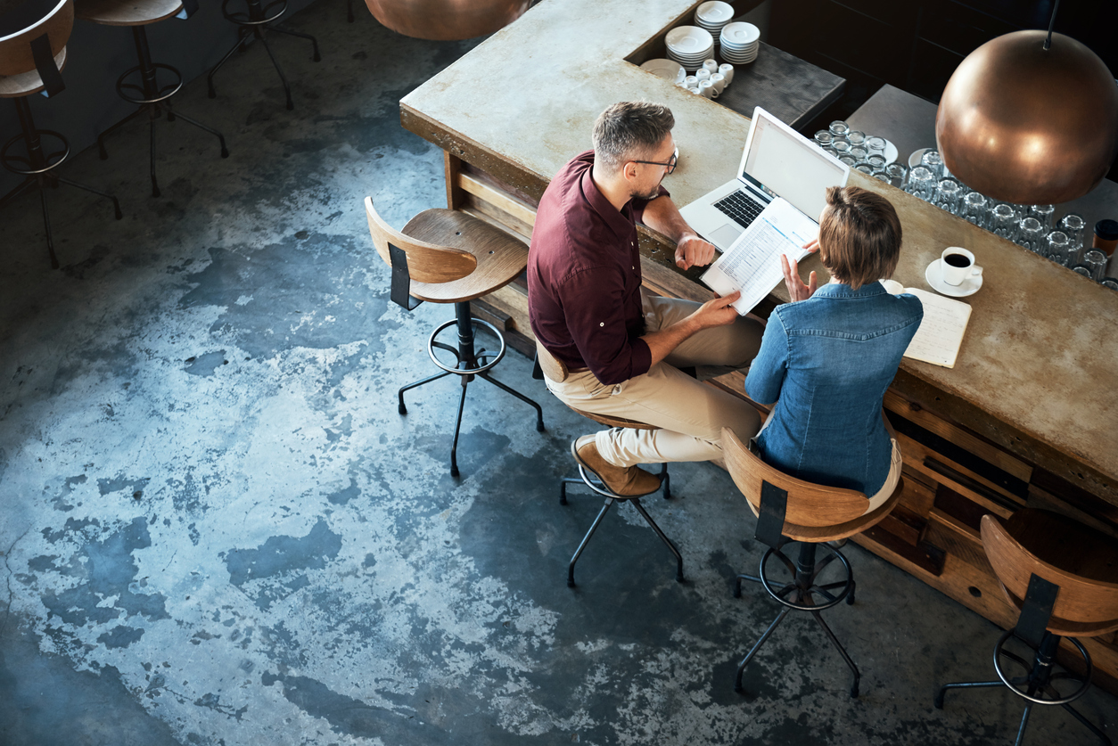 Two business owners sit at their cafe bar discussing metrics over a laptop and stack of papers.