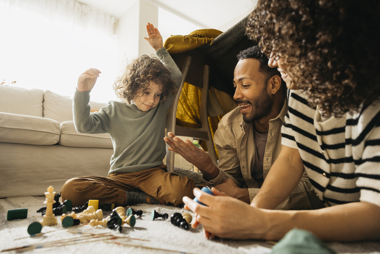 A Mother and Father play chess with their child on their living room floor.