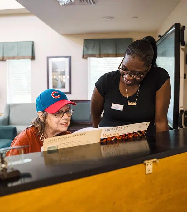 Woman in a red shirt and blue cap engaging with a staff member at a piano, both smiling and discussing music, reflecting person-centered care and connection in a supportive environment.