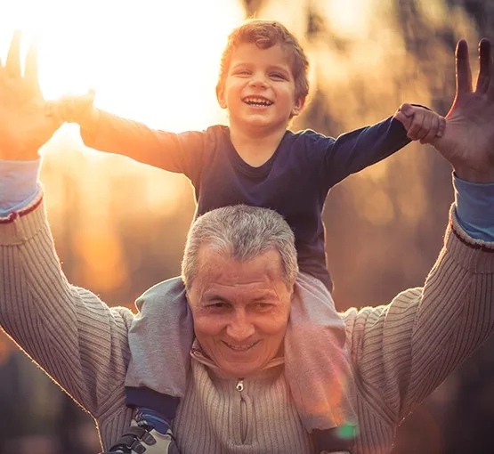 Smiling senior man with gray hair giving a piggyback ride to a joyful young boy, both enjoying a sunny outdoor moment, reflecting the joy and connection emphasized in pet therapy and community support for seniors.
