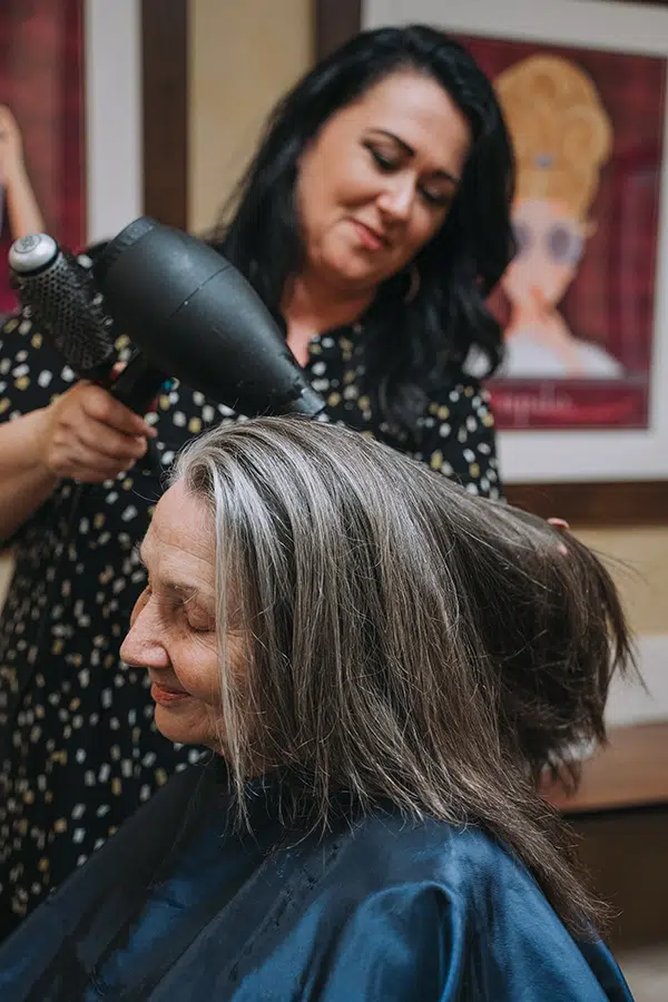 Hair stylist using a blow dryer on a senior woman's hair in a warm, inviting salon setting, reflecting personalized care and attention at Anthem Memory Care.