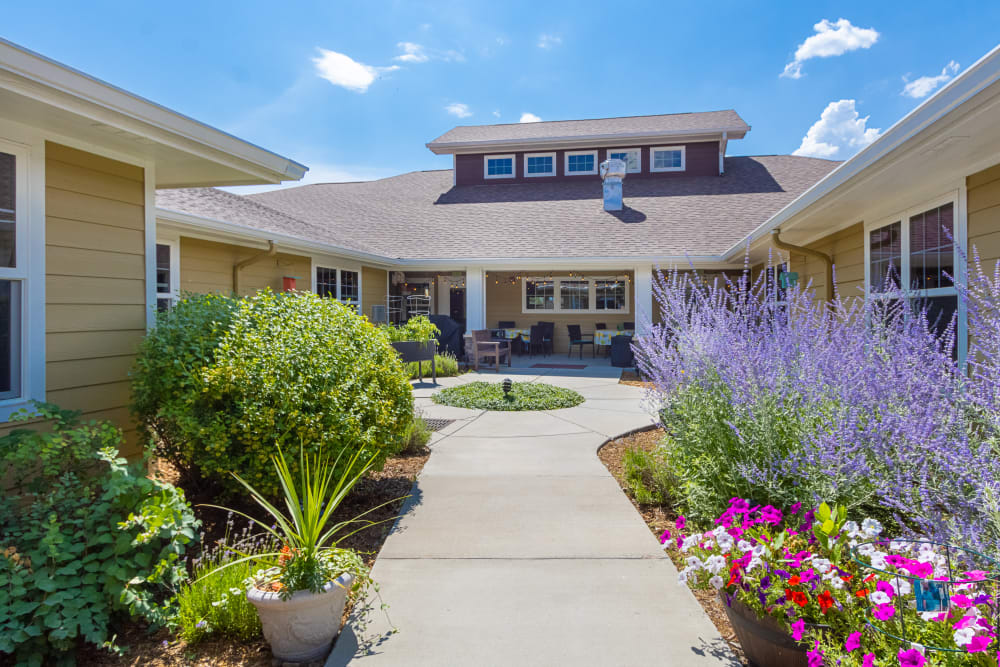 Courtyard of Anthem Memory Care community in Littleton, CO, featuring vibrant flowers, greenery, and a welcoming entrance to the building under a clear blue sky.