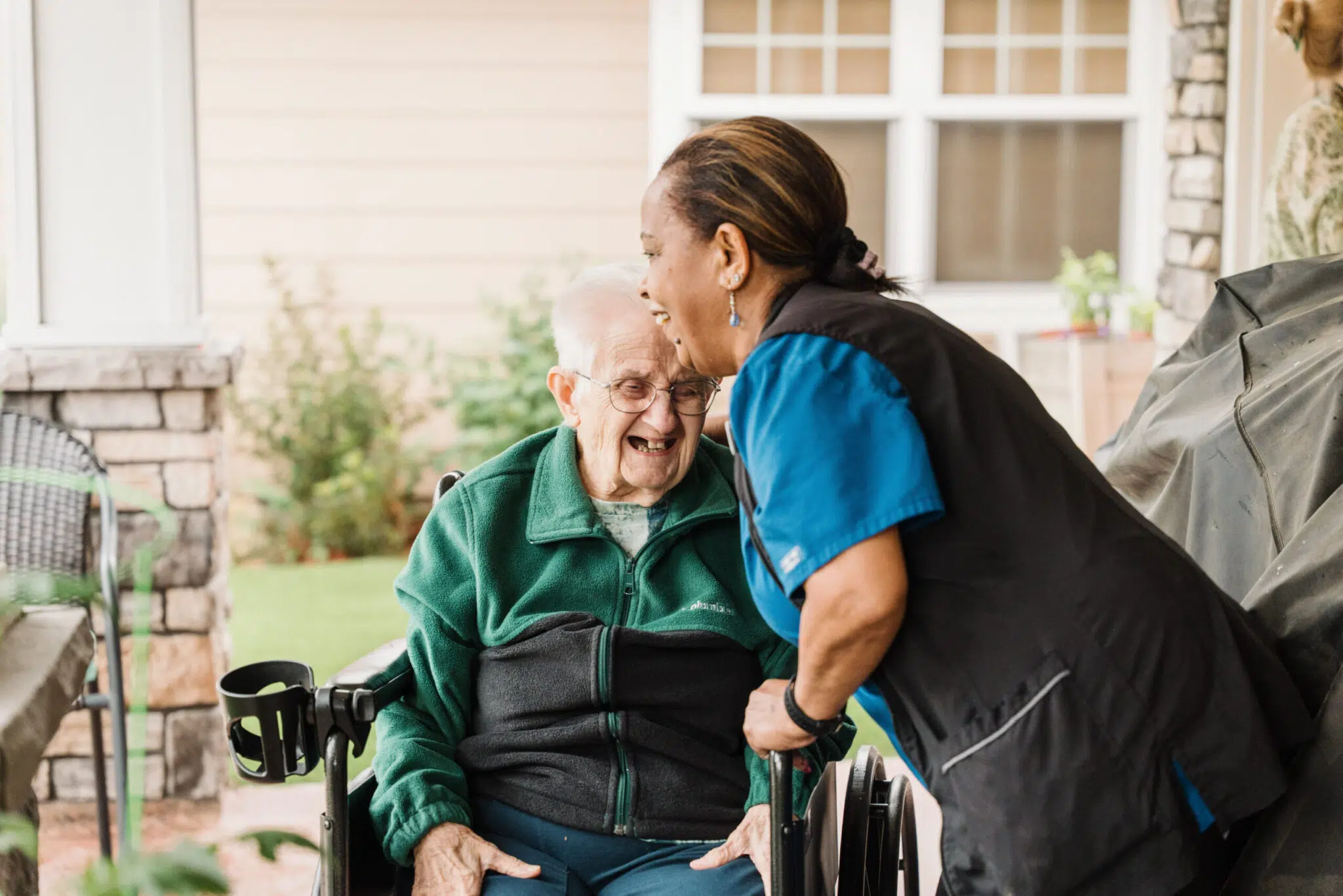 An elderly person in a wheelchair smiles while interacting with a caregiver, both sharing a joyful moment outdoors.