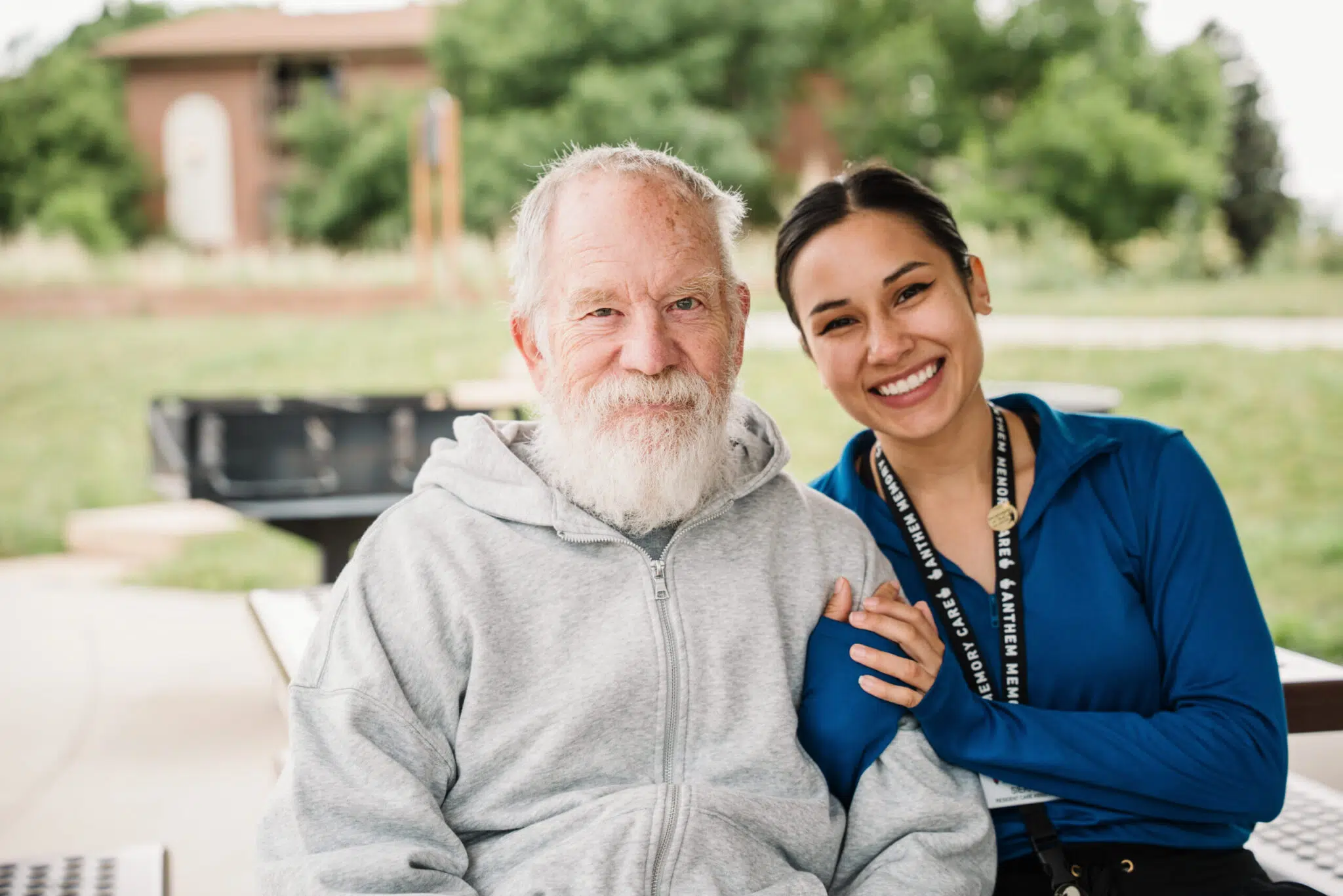 A smiling elderly man with a beard sits next to a young woman, both enjoying a sunny outdoor setting.