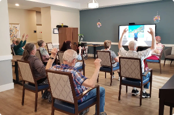 Residents engaging in a group activity at Highline Place Memory Care Community, participating with raised hands in response to a presentation on a screen.