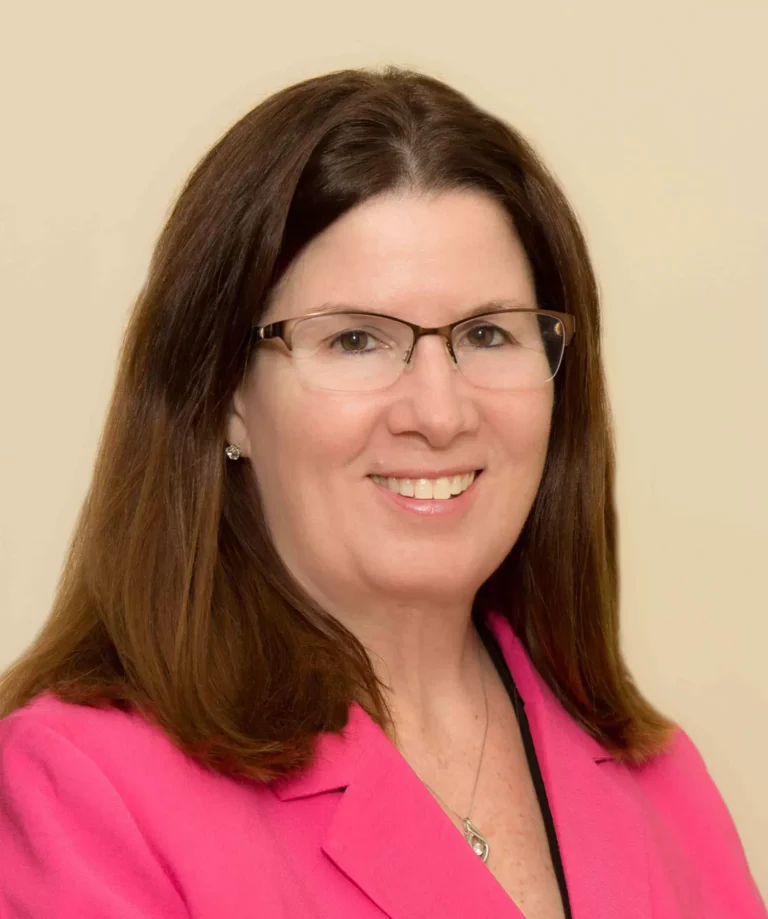 Iva Prinsen, Executive Director at Chelsea Place, smiling in front of a stone wall, wearing a plaid shirt and glasses, showcasing her commitment to compassionate care for seniors with dementia.