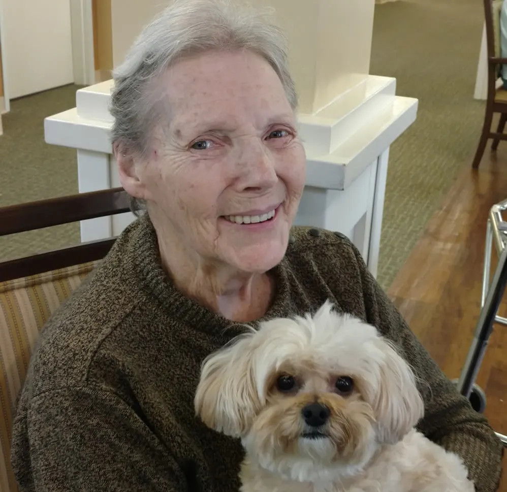 Smiling senior woman holding a small dog, showcasing the joy of pet therapy in a memory care community setting.