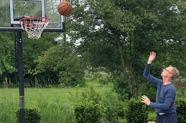 Man shooting a basketball toward a hoop in a green outdoor setting, illustrating recreational activities at Anthem Memory Care community in Tinley Park, IL.