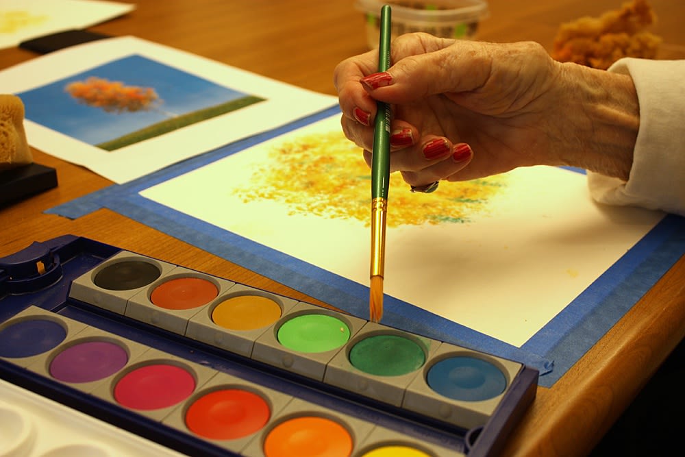 Hand with red nails holding a paintbrush above a watercolor palette, creating art on paper, surrounded by colorful paintings, reflecting creative activities at Chisholm Place memory care community.