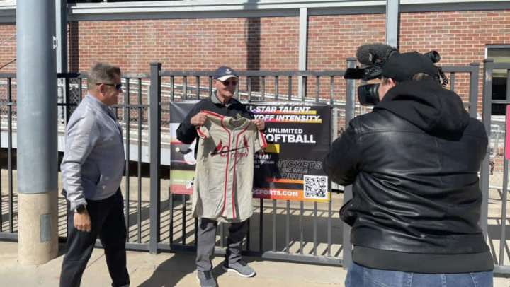 Two men interacting at a baseball venue, one holding a baseball jersey while the other takes a photo, with a promotional banner for softball events in the background, illustrating residents' connection to their passions like baseball.