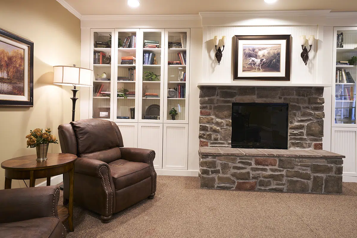 A cozy living room featuring a stone fireplace, bookshelves, a lamp, and comfortable leather chairs.