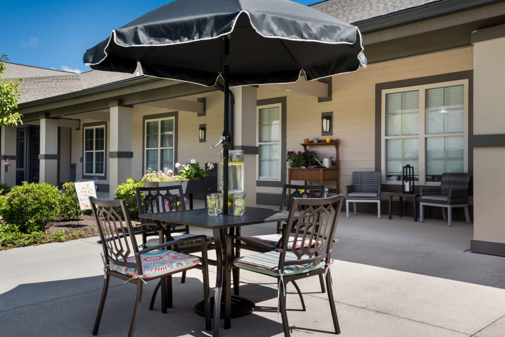 Outdoor seating area at Anthem Memory Care community in West Chester, Ohio, featuring a table with colorful chairs, an umbrella, and landscaped greenery.