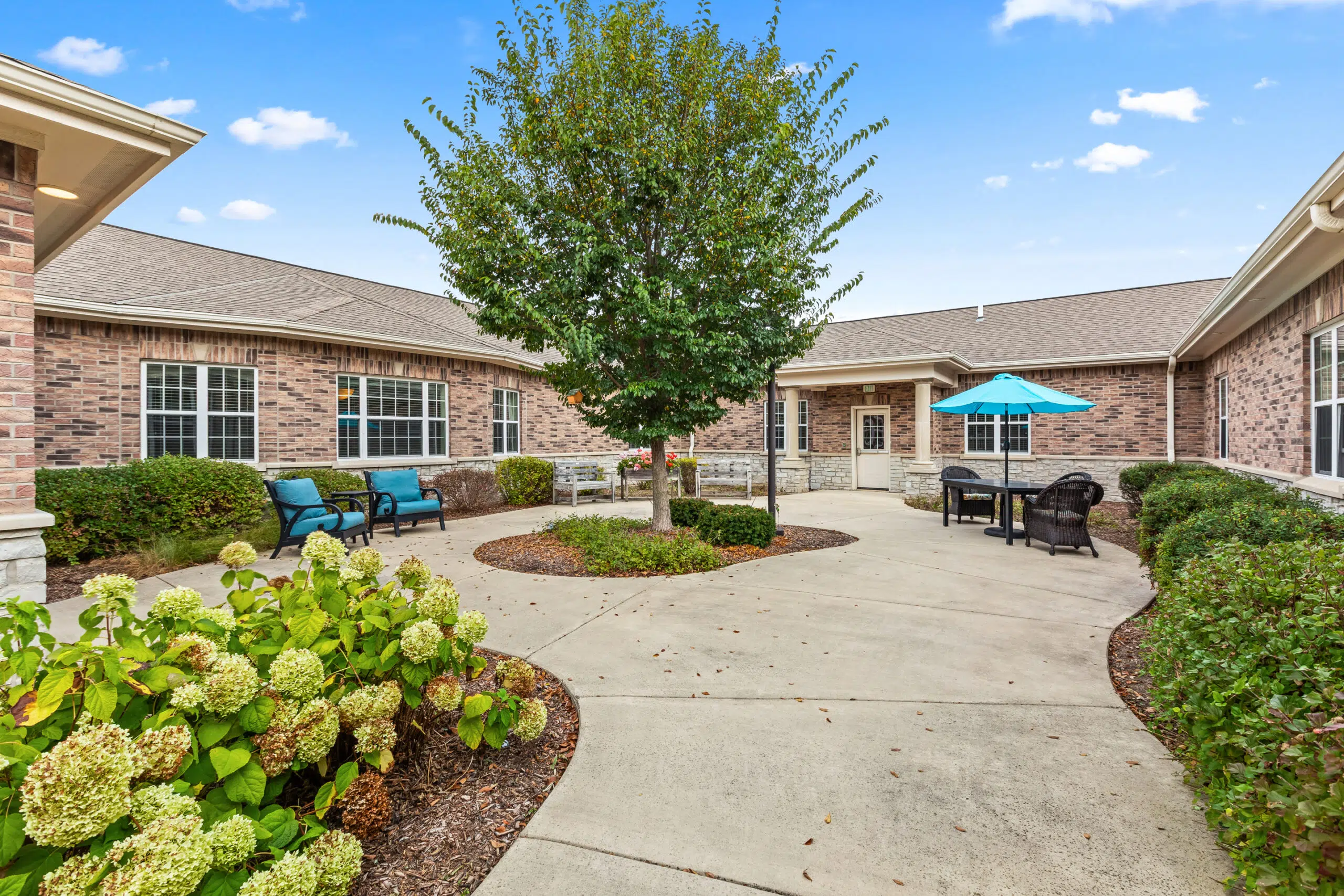 An outdoor courtyard surrounded by the facility buildings, featuring a central paved area, a large tree, teal outdoor seating, and a table with a matching teal umbrella.