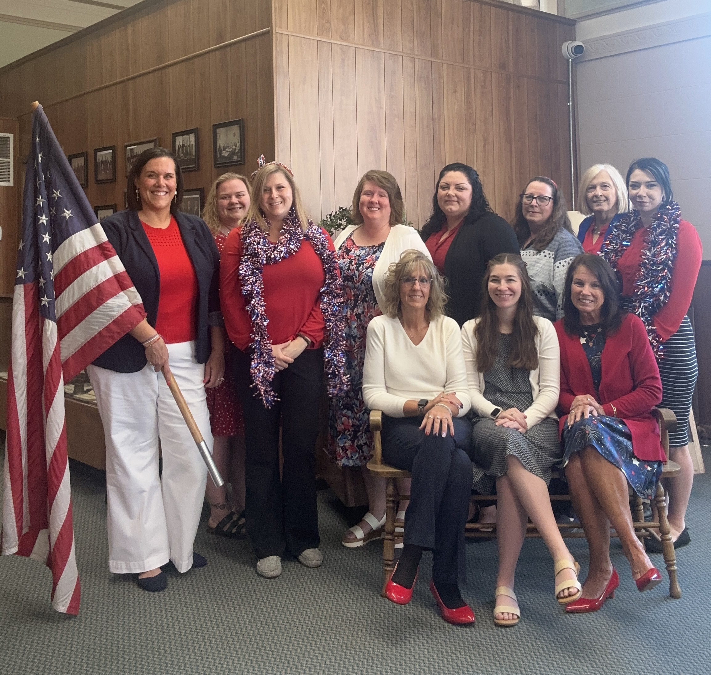 Group of eleven women posing indoors with American flag and festive red, white, and blue attire.
