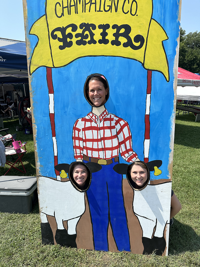 Three women posing with heads through a Champaign Co. Fair cutout of a farmer and two calves.