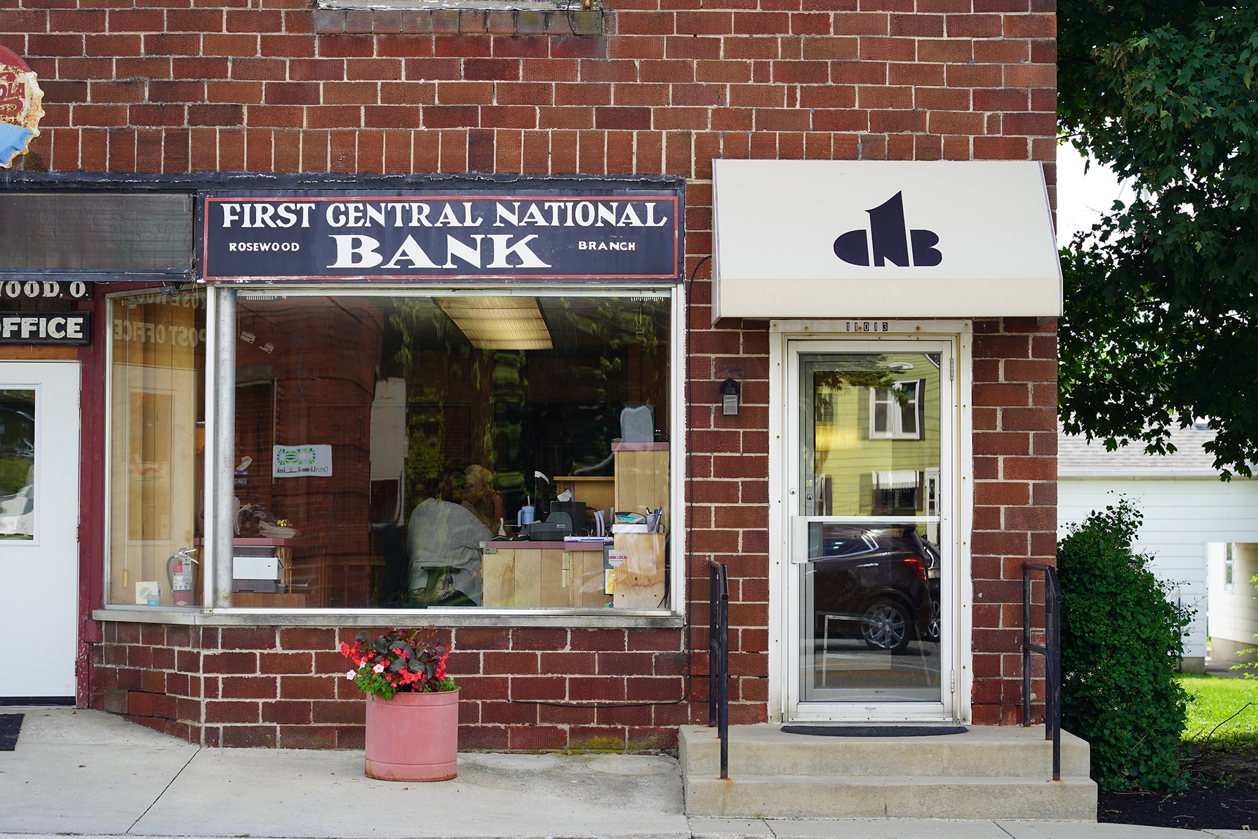 Front of First Central National Bank branch with glass window showing person inside and a small planter with flowers.