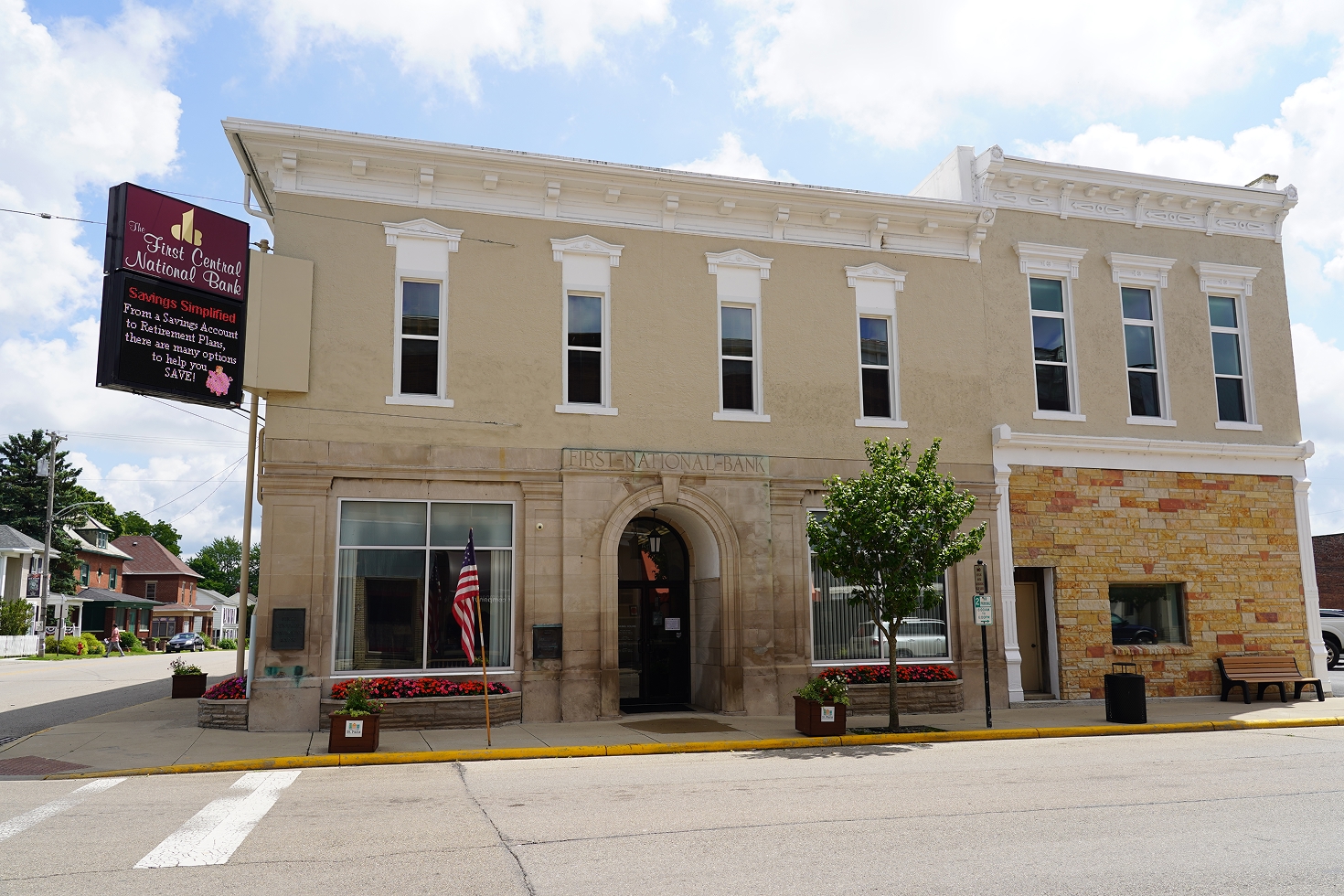 First Central National Bank building with beige facade, arched entrance, flag, and flowers on a street corner.