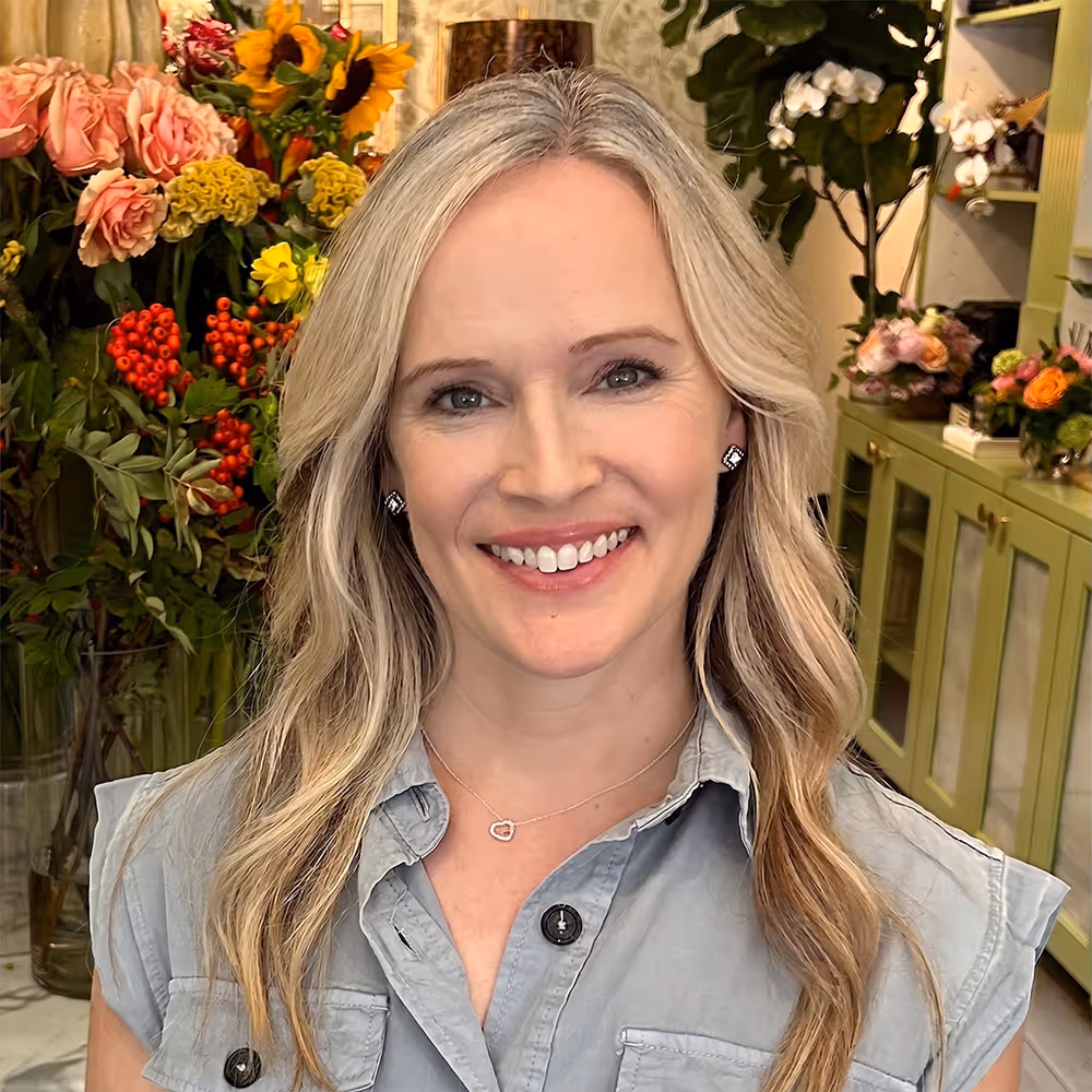 Smiling woman with long blond hair in a sleeveless gray shirt, standing in a room with colorful flowers. Dana Maurice LPC A and owner of Bentley Counseling, PLLC