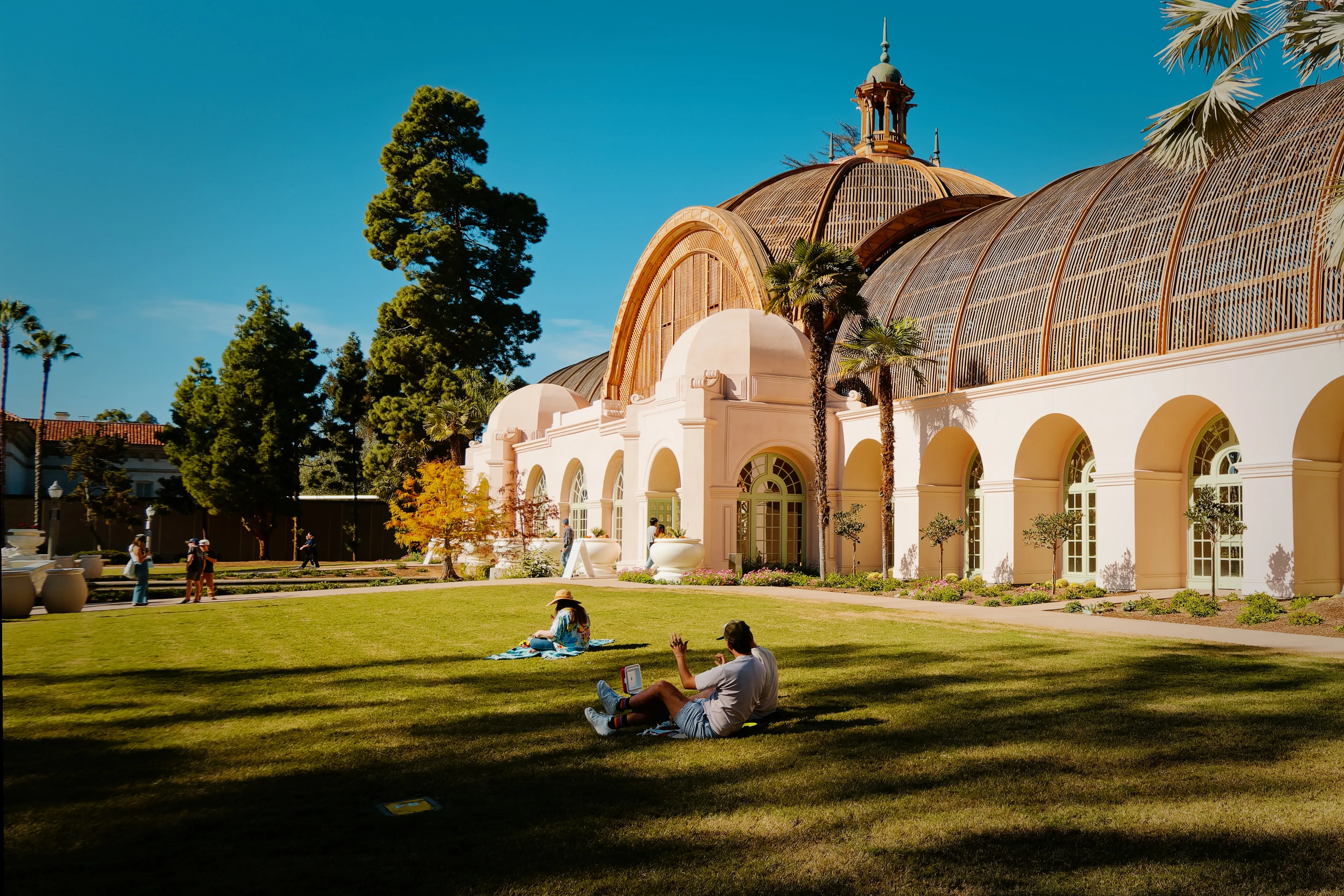 People relaxing on the lawn in front of a historic building with large arched windows and domed roofs at Balboa Park.