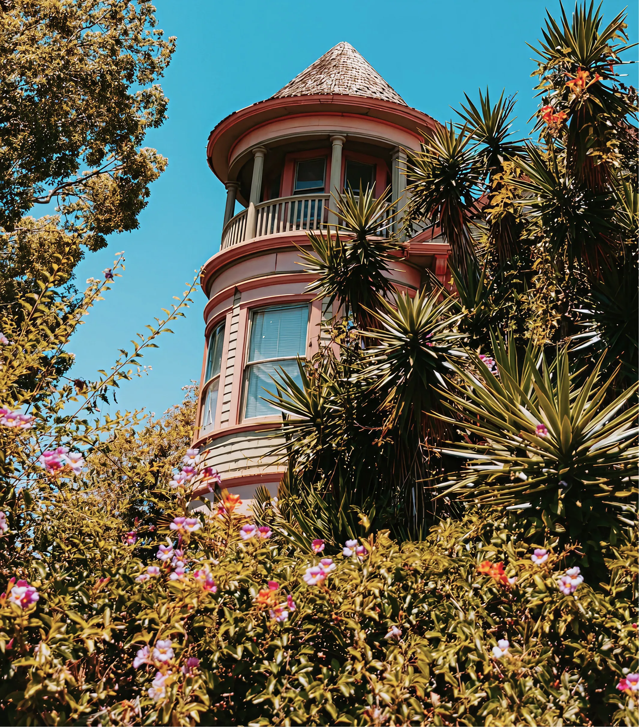 Victorian-style house turret with round windows surrounded by lush green plants and colorful flowers.