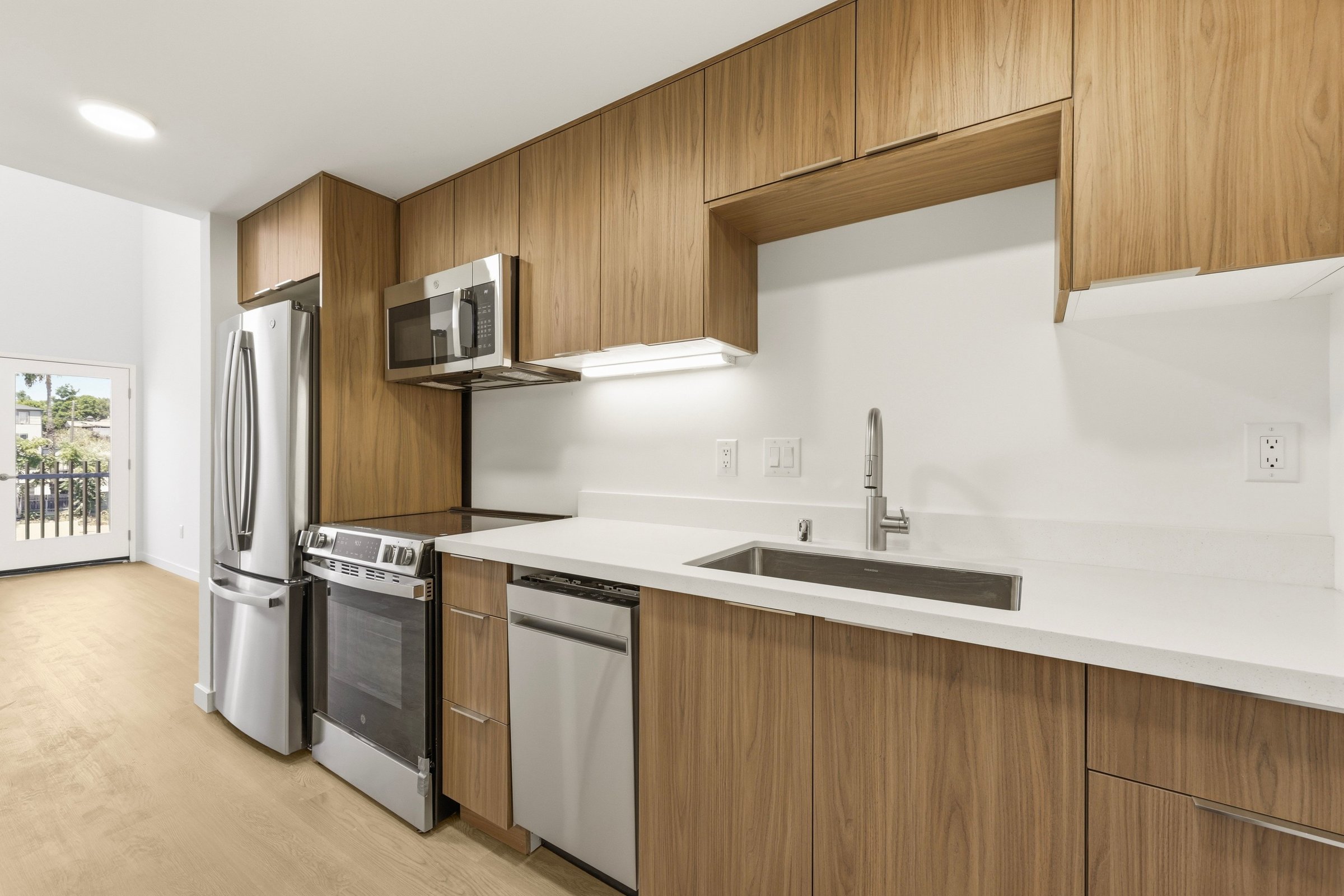 Modern Goldilocks apartment kitchen with warm wood cabinetry, stainless steel appliances, white countertops, and natural light.